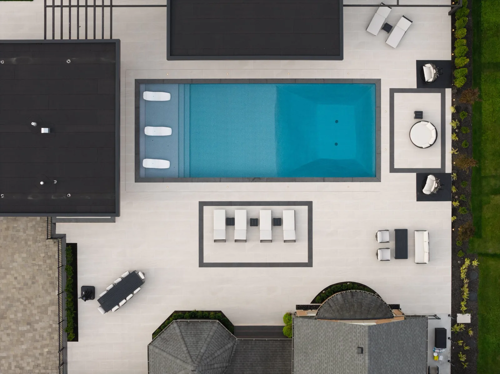 Aerial view of a backyard pool terrace paved in large-format light porcelain, with charcoal-bordered lounge pads, lighting set into the paving and planting along the fence line